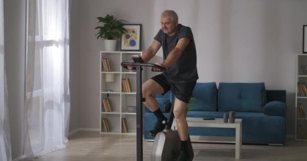 Zapobieganie chorobom układu krążenia w średnim wieku, człowiek trenuje na rowerze stacjonarnym w domu — Wideo stockowe