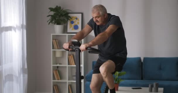 Zmęczony mężczyzna trenuje na rowerze stacjonarnym w domu, średni strzał siwych włosów osoby z wąsami — Wideo stockowe