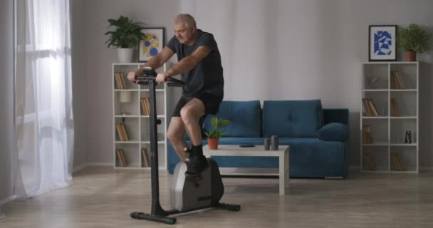 Fitness em casa, homem de meia-idade está treinando em bicicleta de exercício na sala de estar, treino cardio — Vídeo de Stock