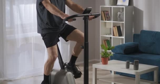 Träning med stationär cykel i hemmiljö, mannen snurrar pedaler i vardagsrummet, workout — Stockvideo