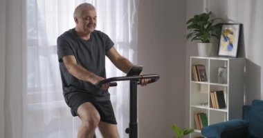 Egzersiz bisikleti üzerinde sabah kardiyo eğitimi, orta yaşlı bir adam hafta sonu oturma odasında spor yapıyor.