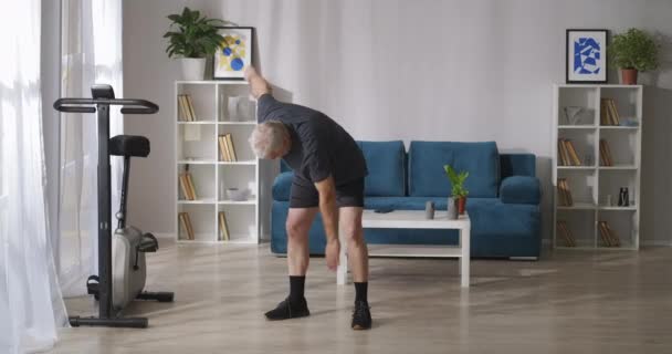 Homem de meia-idade está treinando em casa de manhã, inclina e exercícios de alongamento, ginástica para articulações — Vídeo de Stock