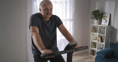 Sağlıklı orta yaşlı bir adam hafta sonu evde bisikletle antrenman yapıyor. Oturma odasında pedal çeviriyor.