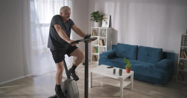 Treino com bicicleta estacionária em casa, homem de meia idade está treinando e girando pedais, mantendo-se em forma e saúde — Vídeo de Stock