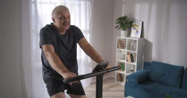 Müder älterer Mann tritt in die Pedale eines stationären Fahrrads, trainiert allein zu Hause und hält körperliche Aktivität aufrecht — Stockvideo