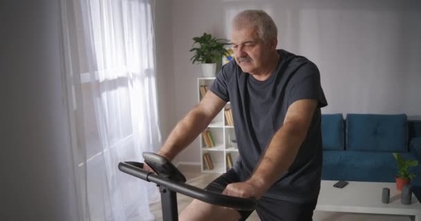 Mann mittleren Alters trainiert zu Hause, tritt in die Pedale des Heimtrainings im Wohnzimmer, lebt gesund und hält körperlich fit — Stockvideo