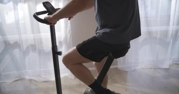 Treinamento em casa com bicicleta estacionária, o homem está girando pedais, mantendo-se em forma durante o auto-isolamento em tempo pandêmico — Vídeo de Stock