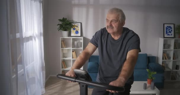 Calm middle-aged man is spinning pedals of exercise bike in home, sport activities with equipment — Stock Video