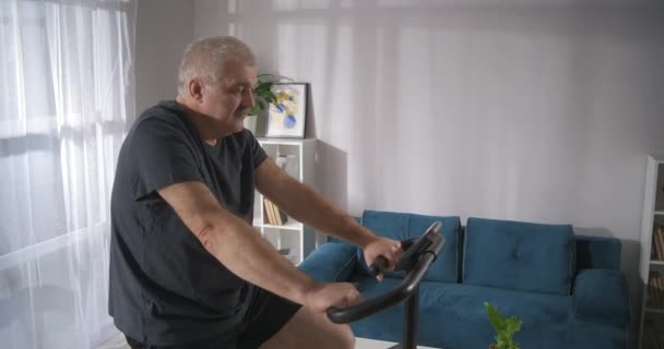 Entraînement avec vélo stationnaire à la maison, homme d'âge moyen est de garder la forme et de perdre du poids par l'entraînement — Video