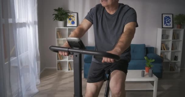 Yetişkin adam evde sabit bisiklet kullanıyor, orta yaşlı insan sağlığa önem veriyor. — Stok video