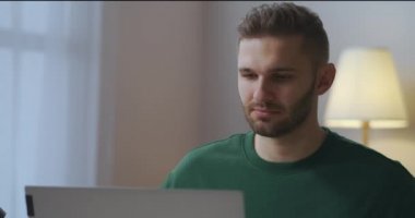 Genç adam evde dizüstü bilgisayarla çalışıyor ve iyi görüş ve koruma için gözlük takıyor.