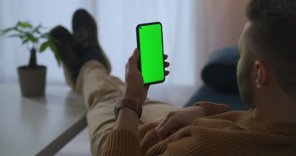 Smartphone mit grünem Bildschirm in den Händen eines entspannten Mannes zu Hause, Benutzer surft im Internet und in sozialen Netzwerken, liest E-Book — Stockvideo