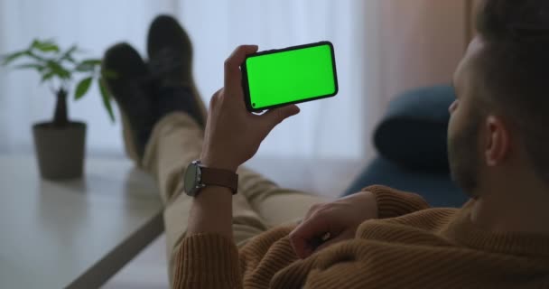 Человек держит смартфон в руке и смотрит видео, отдыхая дома или в офисе, технология зеленого экрана для монтажа — стоковое видео