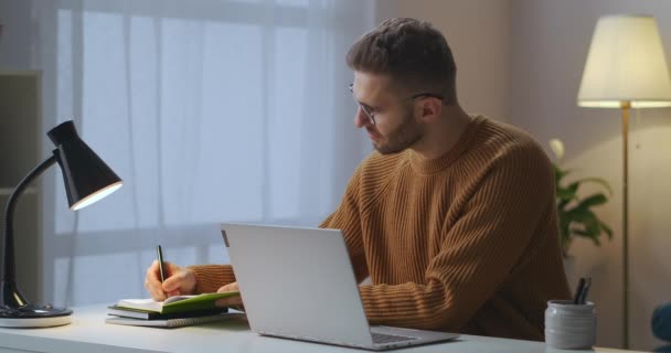 Homem adulto está escrevendo notas no notebook e digitando no laptop, sentado sozinho no quarto, trabalhando remotamente, usando conexão com a internet — Vídeo de Stock