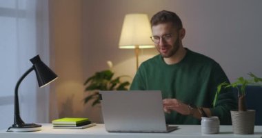 Bilgisayarla çevrimiçi iletişim kuran genç adam, interneti bilgisayardan arayarak akşam evde oturuyor.