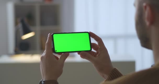 Grön skärm på modern smartphone i händerna på mannen, sitter hemma och tittar på video, närbild visa genom axeln — Stockvideo