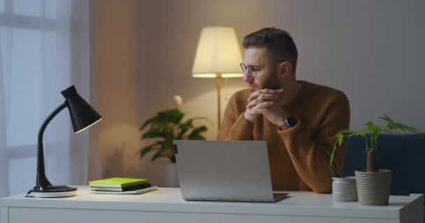 一个戴眼镜的英俊男人正独自坐在房间里，在笔记本电脑前，沉思着，晚上在家里的男性画像 — 图库视频影像