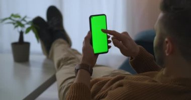 Adam apartmanda dinleniyor, cep telefonu kullanıyor, internette sörf yapıyor ve internetten alışveriş yapıyor, yeşil ekranlı bir alet.