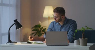 Yetişkin adam dizüstü bilgisayarında ders izliyor ve not defterine not alıyor, internetten öğreniyor, internette bilgi arıyor.