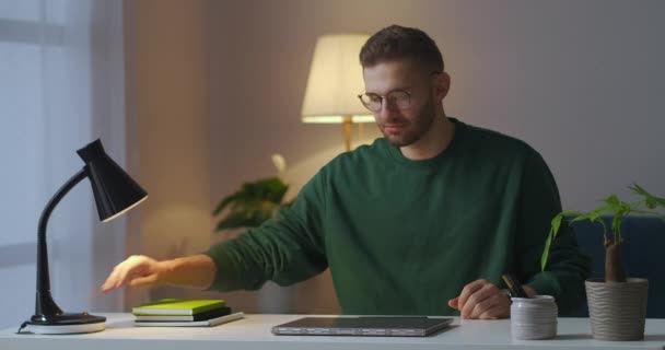 Jongeman met bril gebruikt laptop 's avonds thuis, opent notitieboekje en schakelt tafellamp in — Stockvideo