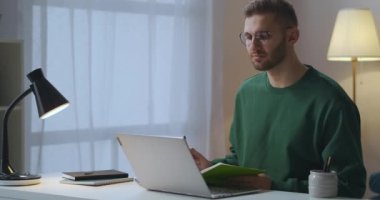 Genç adam online video konferansı sırasında iş arkadaşlarıyla notlar yazıyor, evden çalışıyor, dizüstü bilgisayarla masada oturuyor.