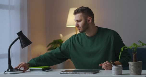 Młody przystojny mężczyzna jest włączanie lampy stołowej i otwieranie laptopa siedzi w salonie w jego mieszkaniu w godzinach wieczornych — Wideo stockowe