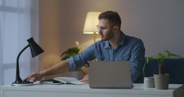 Pohledný student se večer učí online, píše do sešitu a vyhledává informace v notebooku, sedí u stolu v obývacím pokoji — Stock video