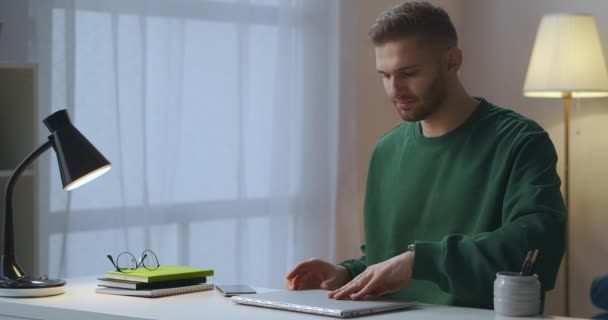 Uomo vestito felpa verde sta digitando sul computer portatile, utente di internet sta lavorando in appartamento, collegandosi alla rete e accendendo lampada da tavolo — Video Stock