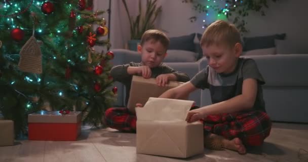 Δύο αγόρια ανοίγουν χριστουγεννιάτικα δώρα καθισμένοι κάτω από ένα στολισμένο χριστουγεννιάτικο δέντρο ανάμεσα σε γιρλάντες — Αρχείο Βίντεο