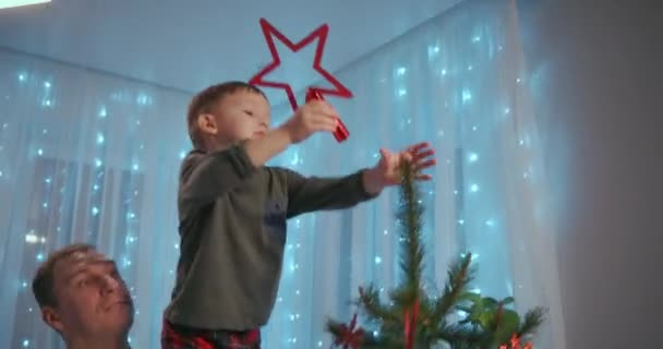 Papá levantó al niño a la parte superior del árbol de Navidad para colocar una estrella de Navidad en la parte superior de la parte superior. Padre e hijo decoran el árbol de Navidad juntos — Vídeos de Stock