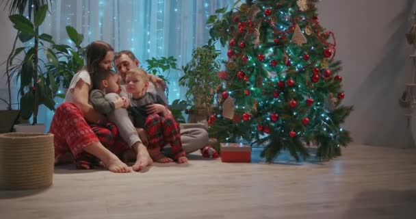 A family with two children sit together at the Christmas tree and cuddle. Love and family happiness on Christmas Eve. Father and two sons cuddle and look at Christmas tree in their living room — Stock Video