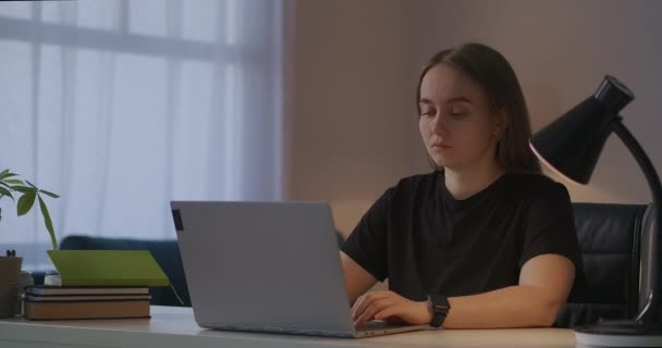 Mujer joven está navegando por Internet por ordenador portátil en casa por la noche, sentado en la mesa y escribiendo en el teclado, la comunicación en las redes sociales — Vídeos de Stock