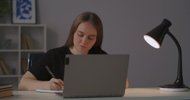 E-learning doma, studentka si prohlíží přednášku online a píše poznámky do sešitu, večer portrét ženy u stolu — Stock video