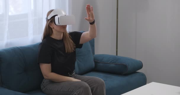 Jovem mulher está usando a cabeça-montado exposição para visualização interior da casa moderna, sentado no sofá na sala de estar — Vídeo de Stock