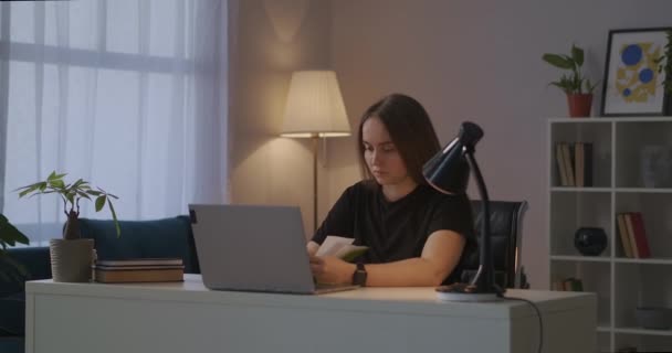 Φοιτητική γυναίκα κάθεται στο τραπέζι με φορητό υπολογιστή και βιβλίο το βράδυ στο σπίτι, μάθηση σε απευθείας σύνδεση, εξ αποστάσεως εκπαίδευση — Αρχείο Βίντεο
