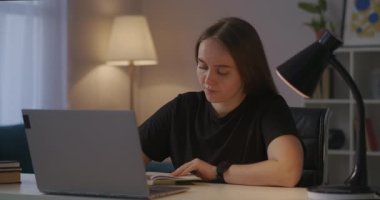 Kadın not defterine not alıyor ve bilgisayar ekranında video dersleri izliyor, akşam evde internetten çalışıyor.