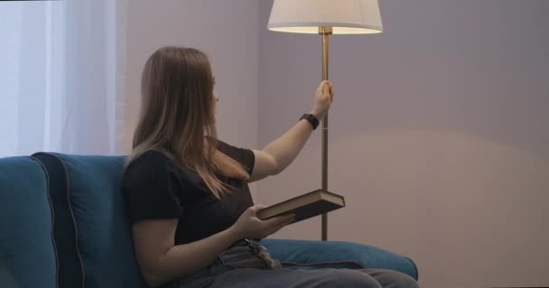 Pretty student woman is reading book at home at evening and switching on floor lamp, rest and relax at weekend — Stock Video