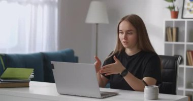 Kadın uzman müşteriye internet üzerinden danışmanlık yapıyor, evinde oturuyor, elinde web kamerasıyla dizüstü bilgisayarının önünde oturuyor ve konuşuyor, el kol hareketi yapıyor.