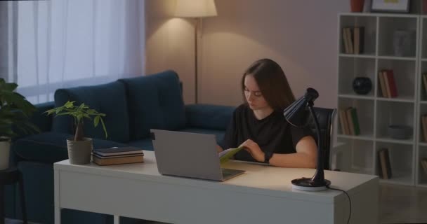 Télétravail de la maison le soir, jeune femme travaille avec un ordinateur portable et prend des notes dans un carnet, assis à table dans le salon — Video