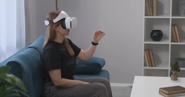 Женщина геймер использует HMD-дисплей, сидя на диване в гостиной, современные технологии виртуальной реальности, женщина жестикулирует руками — стоковое видео