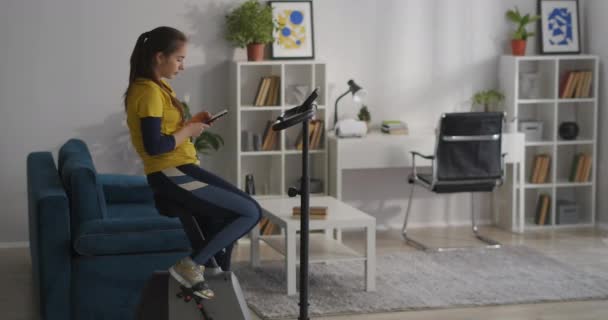 Sportive bavarde dans les réseaux sociaux par téléphone portable pendant l'entraînement avec vélo stationnaire dans le salon, fitness à la maison — Video