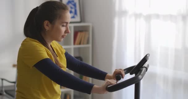 Jeune femme sportive en vélo d'appartement dans le salon, entraînement seul le week-end, équipement sportif pour la remise en forme à domicile — Video