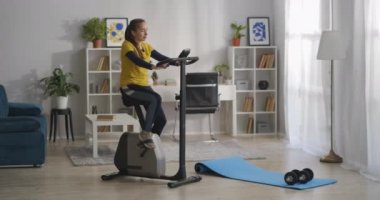 Spor malzemeleriyle oturma odasının tam boy görüntüsü. Genç kadın egzersiz bisikleti, egzersiz ve sağlıklı yaşam tarzı eğitimi alıyor.