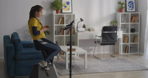 Ung sportig kvinna vilar under träning med cykel, med sociala nät i smartphone, träningsredskap för hemmaträning — Stockvideo