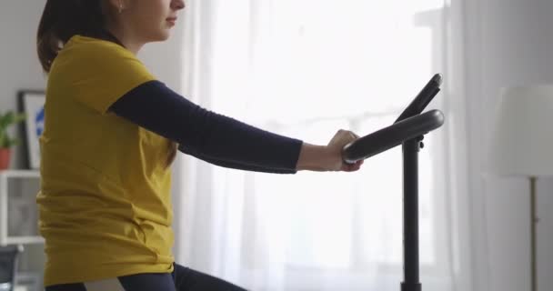 Тренировка с стационарным велосипедом в доме, женщина крутит педали, сохраняя форму и похудение, стройное женское тело — стоковое видео