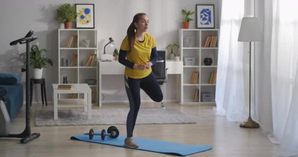 Menina adolescente está fazendo aeróbica na sala de estar de manhã, pisando e levantando a perna no ritmo da música, exercício esportivo engraçado — Vídeo de Stock