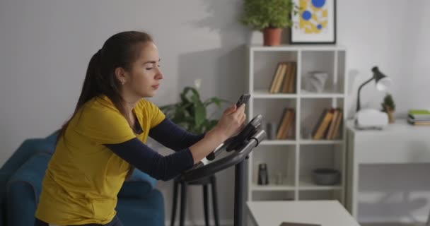Brunette vrouw surft internet via smartphone tijdens het sporten met stationaire fiets thuis, sociale netto verslaving van moderne jongeren — Stockvideo