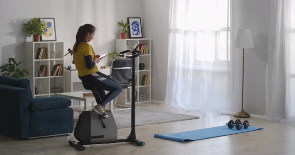 Vrouw typt en verstuurt berichten per mobiele telefoon, zit op hometrainer in de woonkamer van haar flat, pauze in de ochtend workout — Stockvideo