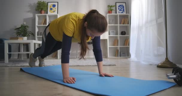 Молодая женщина делает физические упражнения дома, тренировки в квартире в выходные напрягая мышцы ног и рук — стоковое видео