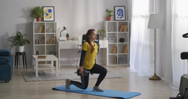 Jovem está fazendo agachamentos com halteres nas mãos, treinando sozinho na sala de estar do apartamento moderno — Vídeo de Stock